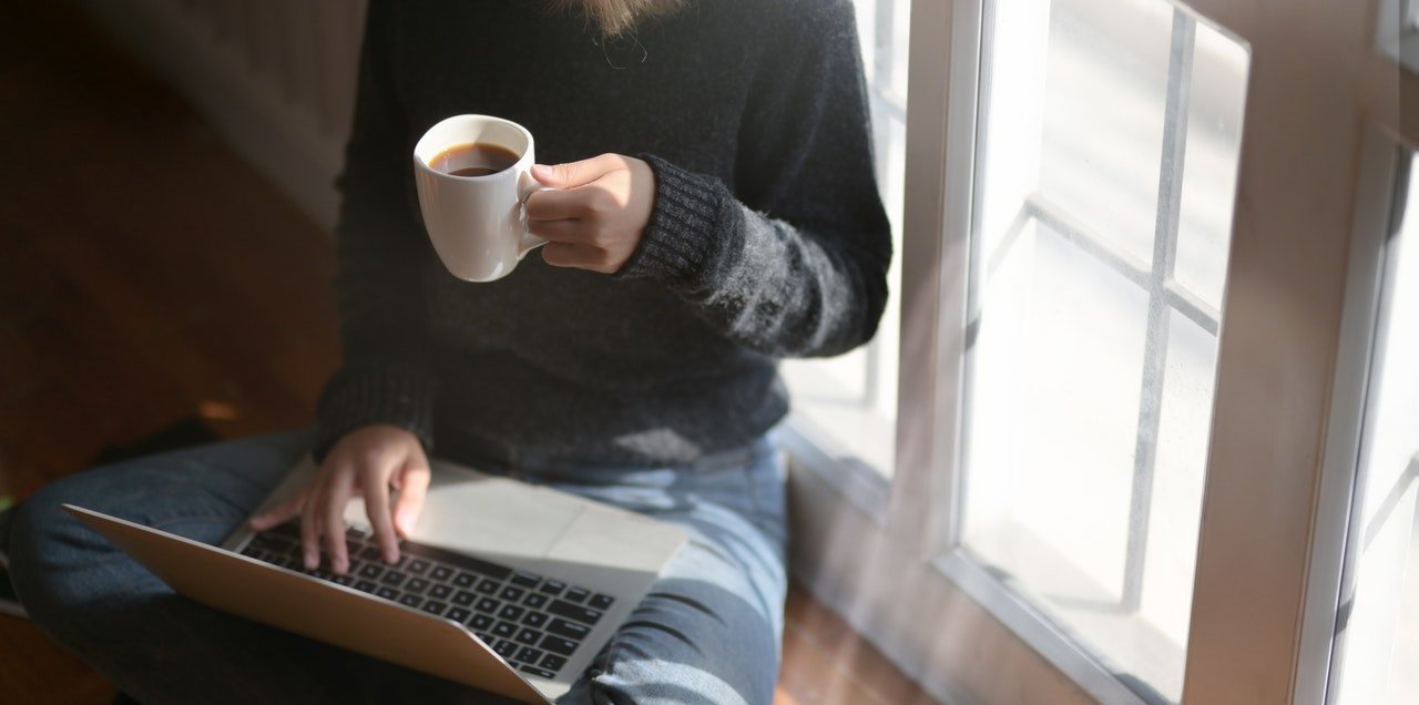A remote worker sitting cross legged on the floor at home with a laptop on their lap and a cup of coffee in their hand